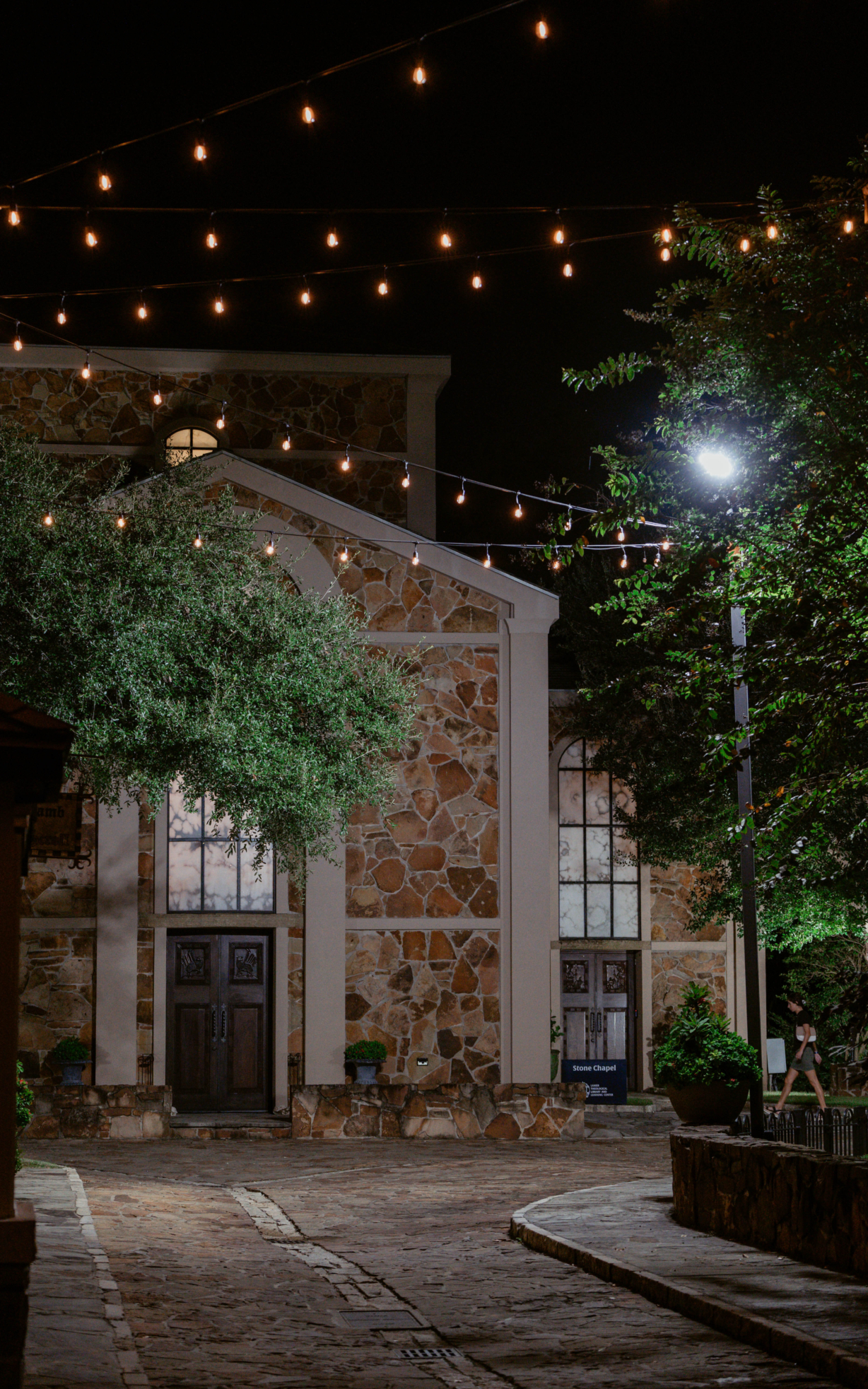 The front of the chapel at night. String lights hang between rows of buildings lining either side of a stone walkway leading to the chapel.