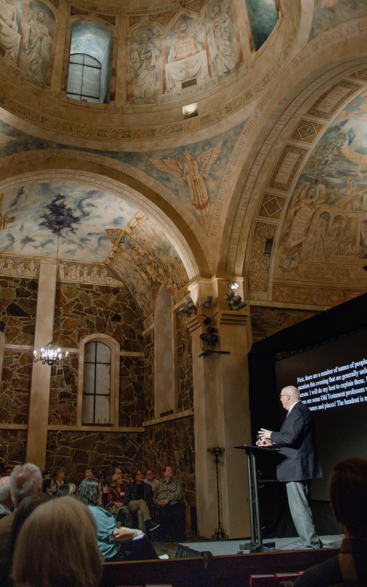 A man speaking to a crowd on stage inside the chapel. The ceiling is painted with images depicting stories from the Bible.