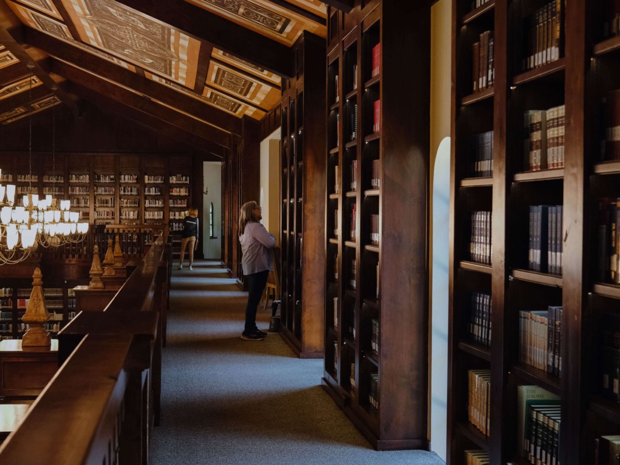 A woman searching for a book on one of the shelves. Sun streams in from the windows.