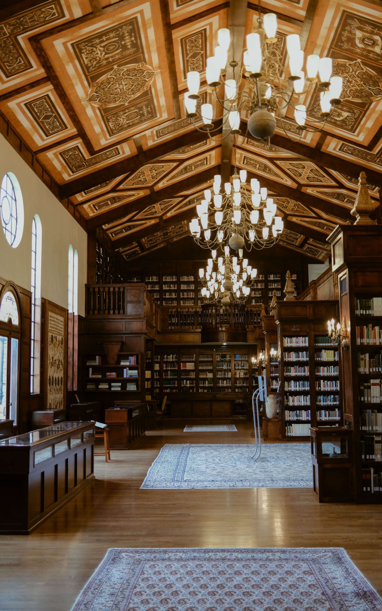 Inside the library. The ceiling is two stories high and hand painted. Three large chandeliers hang from the ceiling and reflect off of the wooden floor. Several rugs line the walkway. Shelves are full of books and there are display cases with artifacts in them.
