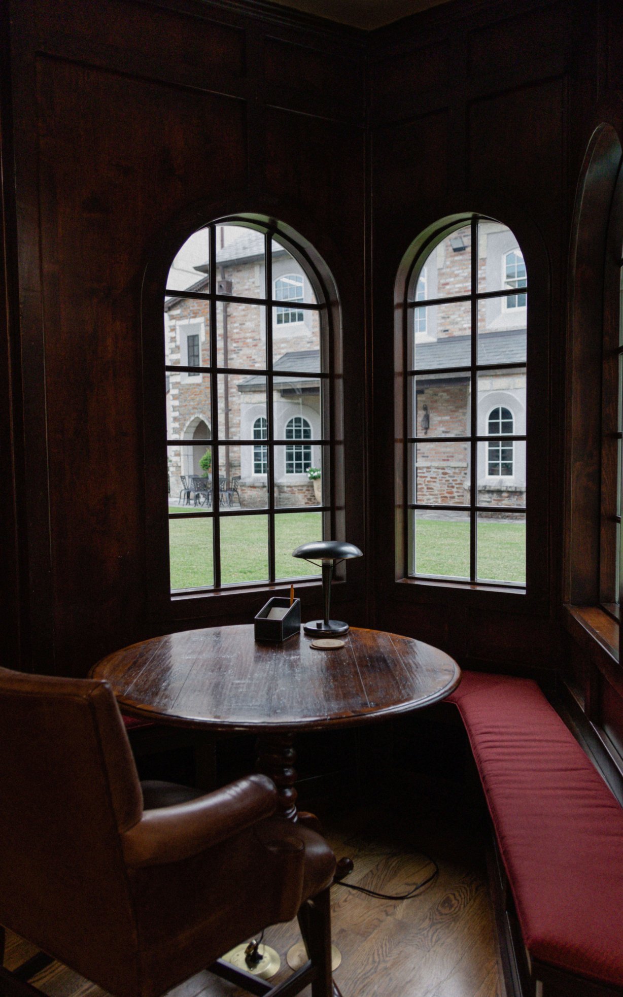 A small round table and chair situated in an alcove of three windows. Under the windows is a bench wrapping around the table. The library courtyard can be seen through the windows.